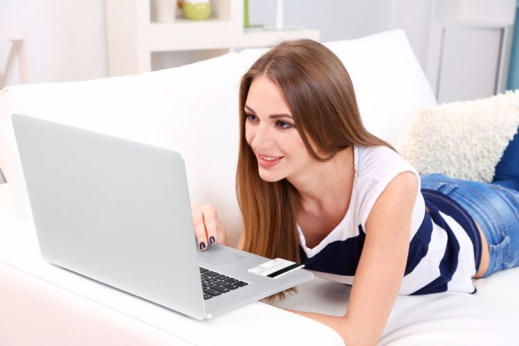 Młoda kobieta siedzi z laptopem na kanapie i trzymając kartę kredytową w dłoni, w domu